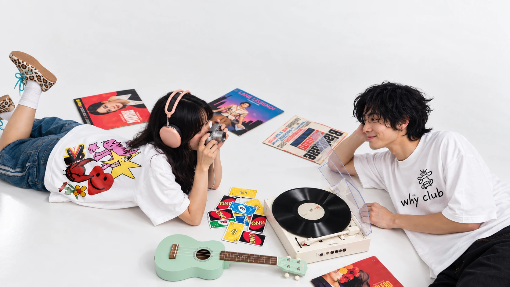 WhÿClub models on a white studio floor with vinyl, camera, and graphic tees.